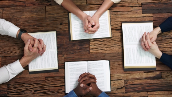 Aerial view of four hands folded on open Bibles and gathered around a table