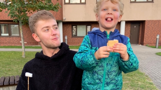 Pavlo sits next to and smiles at the Bronsink's young song while holding a s'more