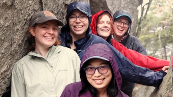 Rachel Kodde and friends pose from Act 5 wearing rain jackets and posing for a photo in front of a boulder