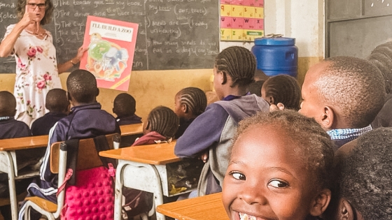 A child smiles in a classroom while Carolyn reads from a book