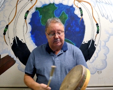 A man beating a drum and explaining aspects of Cree culture